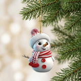 A white acrylic snowman Christmas ornament with red buttons and a striped scarf hangs among green branches against a blurred cream background.