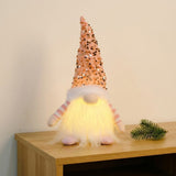 A Christmas gnome with a tall pink sequined hat and glowing white beard stands on a wooden cabinet beside an evergreen sprig.