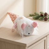 A pink Christmas gnome lies on a wooden cabinet, revealing its circular white base, battery compartment cover, and black switch.