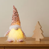 A Christmas gnome with a pink sequined hat and glowing white beard stands beside a wooden tree ornament against a beige wall.