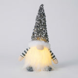 A Christmas gnome with a gray sequined hat, striped sleeves, gray shoes and glowing white beard stands against a light gray background.