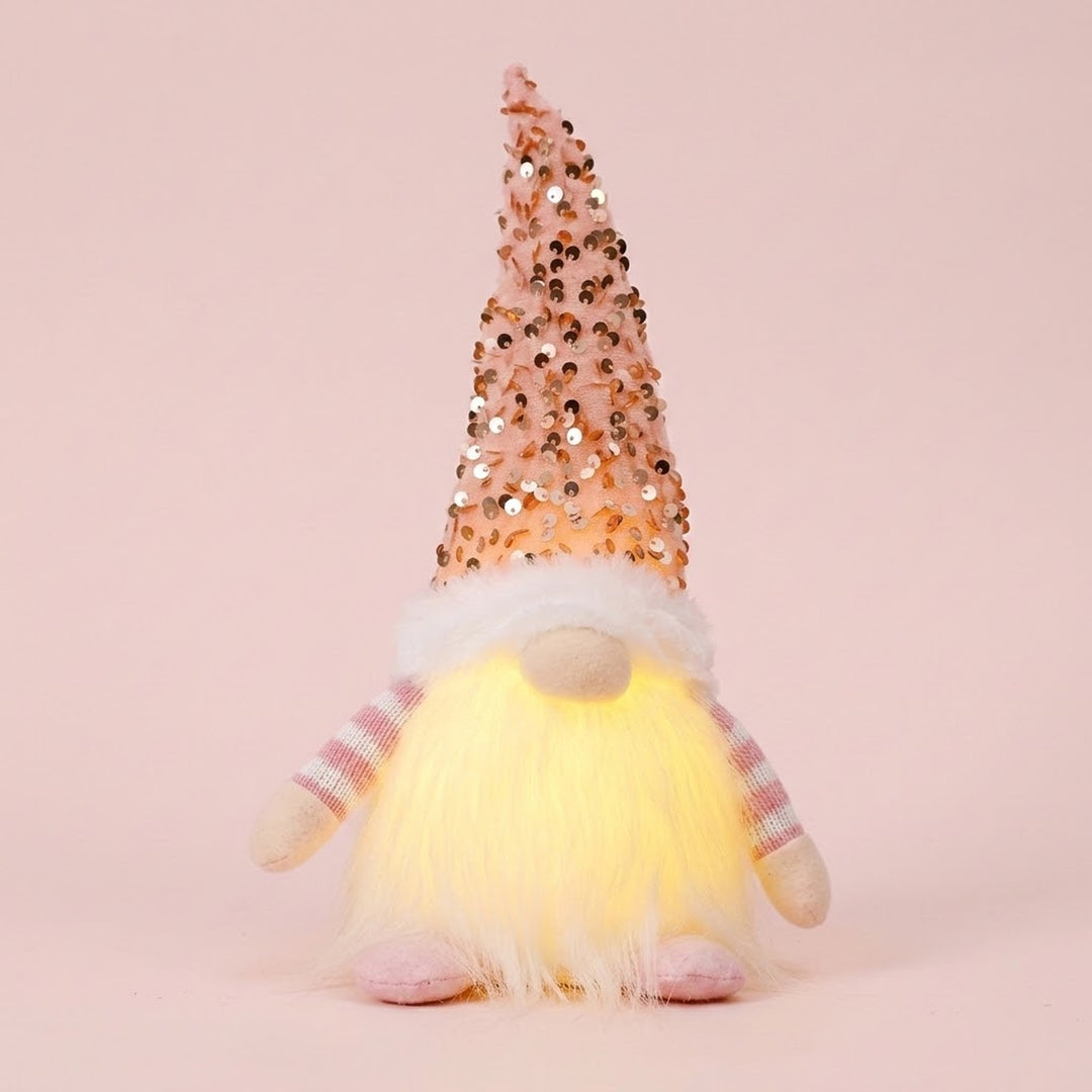Glowing Christmas gnome with a pink sequin hat, white beard, plaid sleeves and pink shoes stands against a pale pink background.