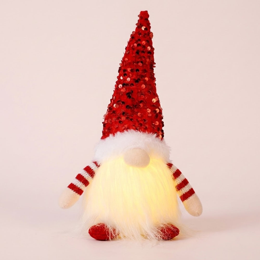 Glowing Christmas gnome with a red sequin hat, white beard, striped sleeves and red shoes stands against a pale pink background.