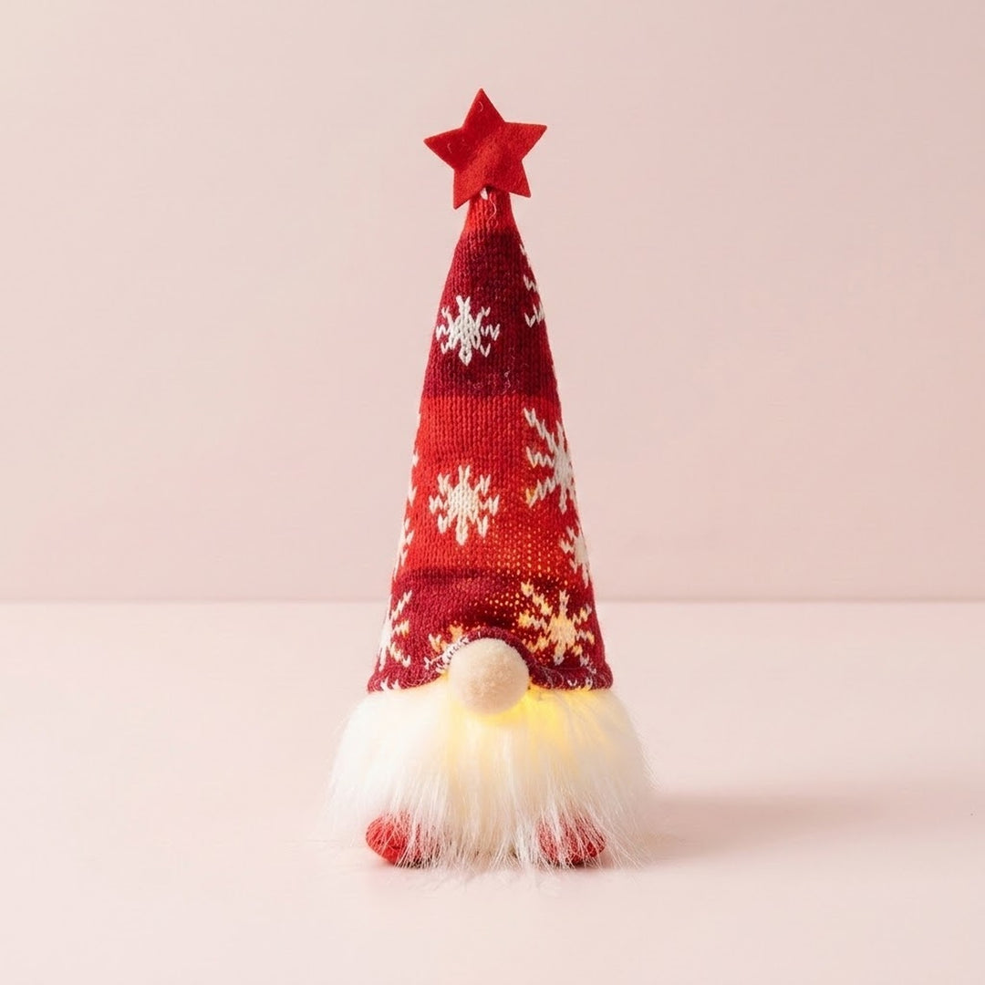 A Christmas gnome with a red knitted snowflake hat, star topper and glowing white beard stands against a pale pink background.