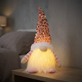 A Christmas gnome with a tall pink sequined hat and yellow-lit white beard stands on a wooden bedside table in a dim bedroom.