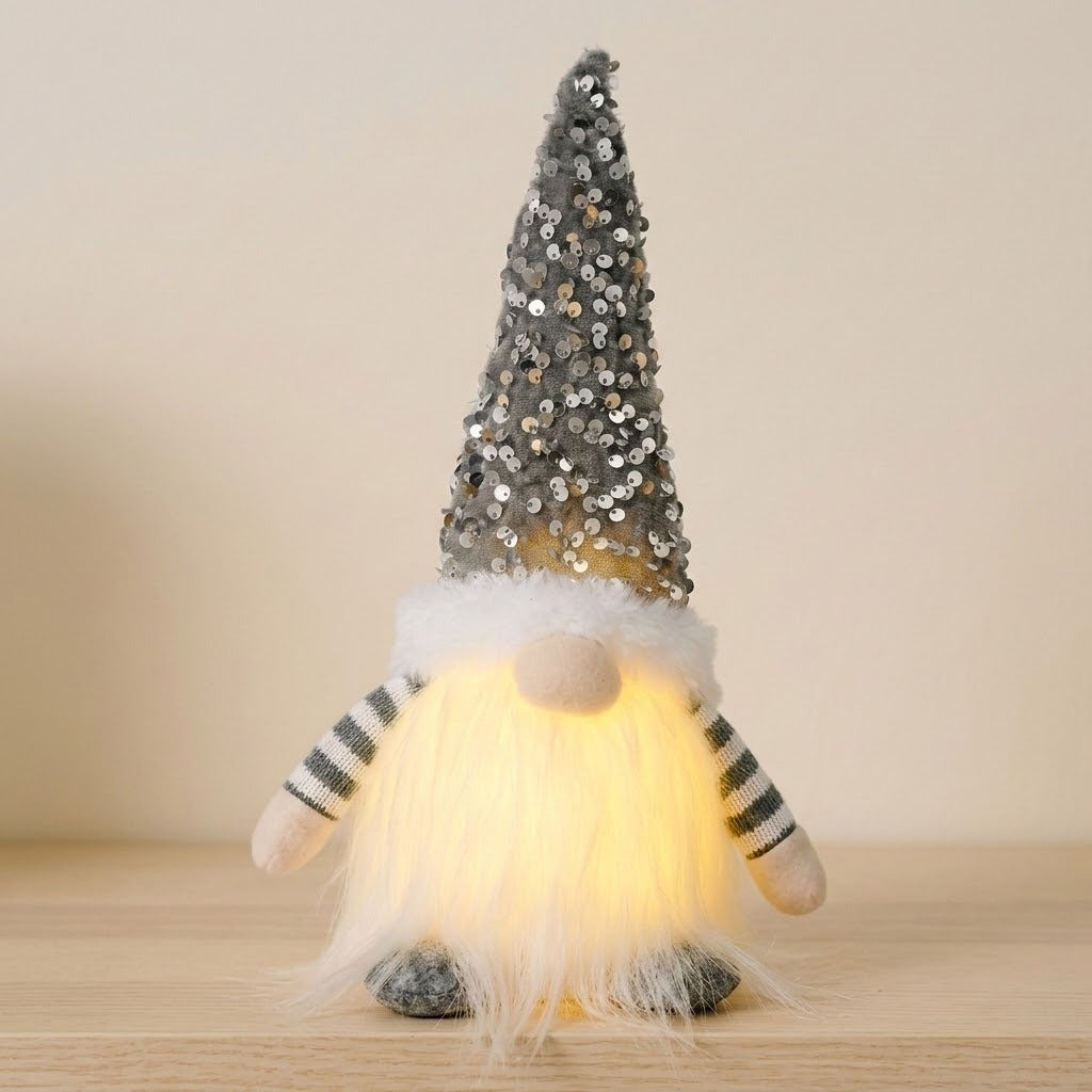 A Christmas gnome with a gray sequined hat, striped sleeves and glowing white beard stands on wood against a beige wall.
