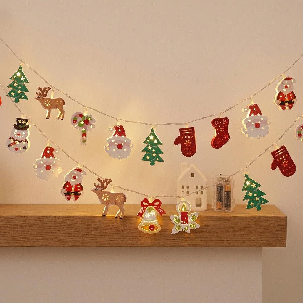 Warm glowing Christmas string lights with colorful festive cutouts hang above a wooden shelf holding a white miniature house.