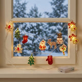 A glowing string light with colorful Christmas ornaments drapes across a white window overlooking snowy trees, with a clear battery box on the sill.