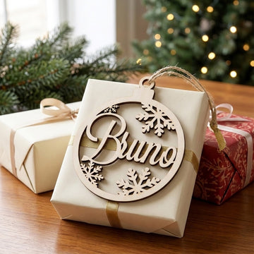 Pale wooden Bruno snowflake ornament rests against a cream gift with gold ribbon, surrounded by presents and blurred Christmas tree lights.
