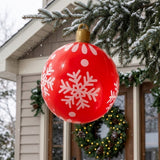 A red inflatable ornament with white snowflakes and a gold top hangs from a snowy evergreen branch near a wreath-decorated door.