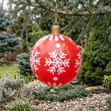 A red inflatable ornament with white snowflakes and a gold top hangs by twine from an evergreen branch above a pebble garden.