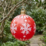 A red inflatable ornament with white snowflakes and a gold top hangs from a tree branch beside a leafy garden path.