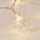 Clear plastic snowflake string lights glowing with a warm white light, shown close-up against a soft beige background.