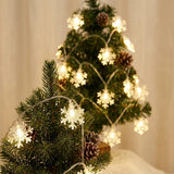 Warm white snowflake string lights wrap two small green Christmas trees with brown pinecones against a cream background.
