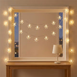 Warm white snowflake string lights drape across and around a white window frame above a wooden table with a small lamp.