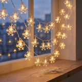 Warm white snowflake-shaped string lights draped across a window ledge, glowing softly in a cozy indoor setting with a city view outside.