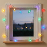 Multicolored snowflake string lights frame a white window above a wooden sill, with a blurred evening view outside.