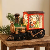 Red and green plastic Christmas train decoration with an illuminated Santa scene sits on a rustic wooden table beside evergreen branches.