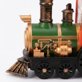 Christmas train decoration close-up shows a green boiler, gold-colored pipes, bronze-colored chimney, and black spoked wheels against a white background.