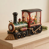 Green and red Christmas train decoration with gold detailing on a light wooden surface, its lit cab showing Santa and a tree.