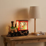 Lighted red and green Christmas train decoration on a rustic wooden table beside a beige table lamp.
