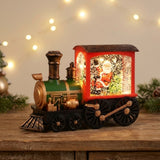 Red and green Christmas train decoration with a glowing Santa scene sits on a wooden cabinet beneath blurred string lights, beside evergreen sprigs.