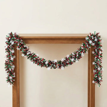 Red and green tinsel garland with white snowflakes drapes across a wooden frame against a cream wall.