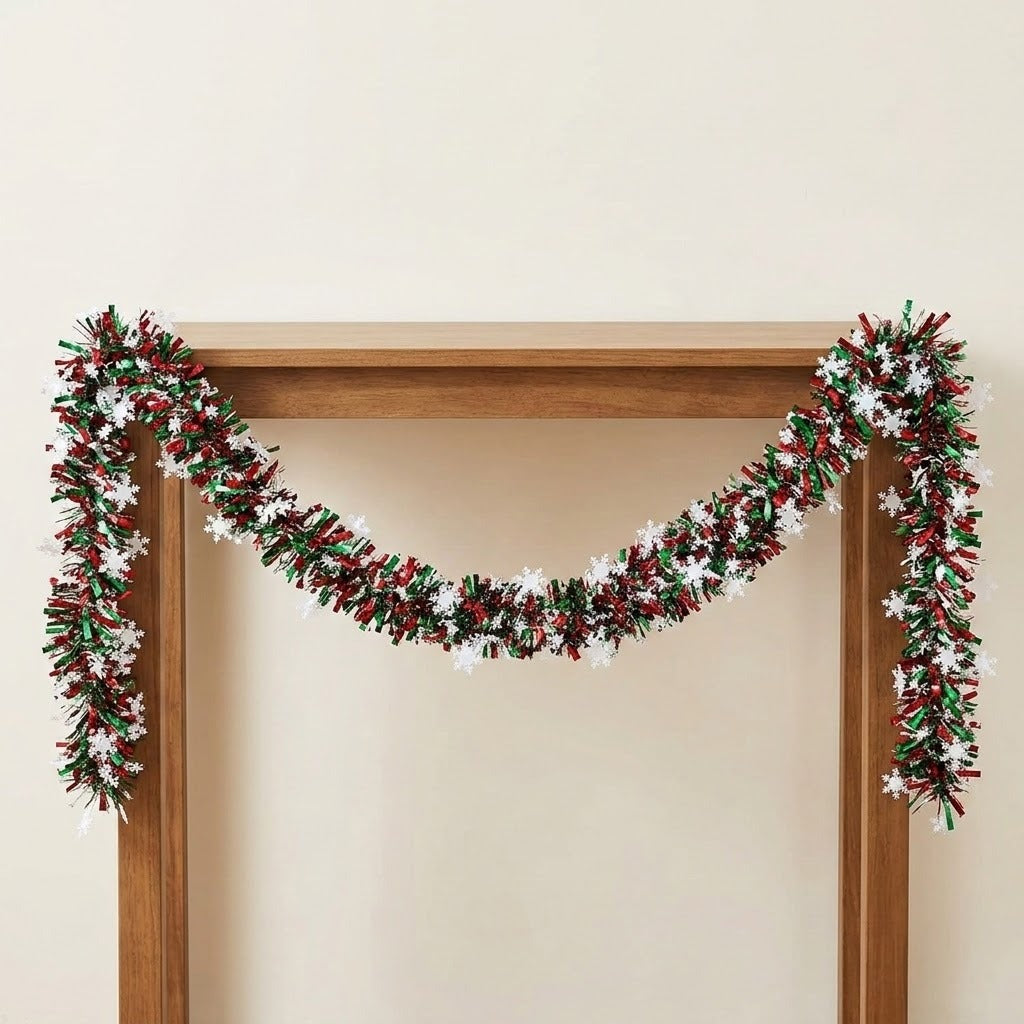 Red and green tinsel garland with white snowflakes drapes across a wooden frame against a cream wall.