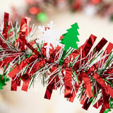 Close-up of red, green, and silver metallic tinsel garland with white snowflake and green tree cutouts against a blurred cream background.