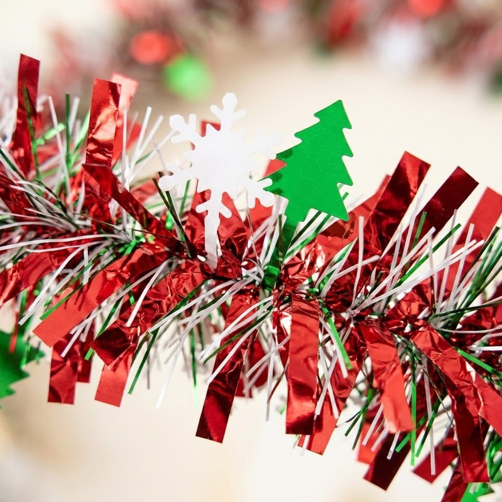Close-up of red, green, and silver metallic tinsel garland with white snowflake and green tree cutouts against a blurred cream background.