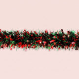 Red and green tinsel garland with wide reflective strips runs horizontally across a pale pink background.