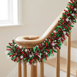 Red and green tinsel garland with white snowflakes follows a curved wooden stair railing beside a window.