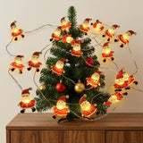 Warm glowing red Santa string lights wrap around a small Christmas tree with red and gold baubles on a wooden cabinet.