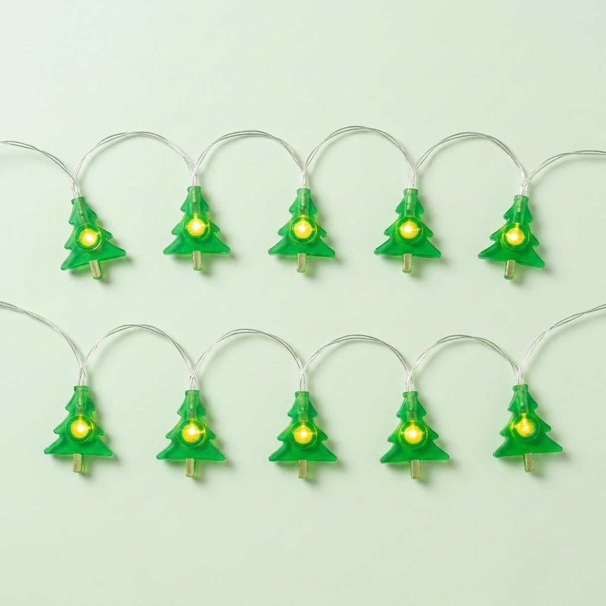Green Christmas tree string lights with glowing yellow centers and silver wires form two rows against a pale green background.