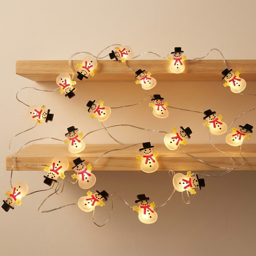 Glowing white snowman string lights with black hats and red scarves drape across two wooden shelves against a cream wall.