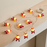 Red Santa string lights glow along a pale wooden ledge, with three figures hanging over the edge and wire coiled above.