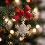 A red-bow snowflake drop earring with clear rhinestones and gold-colored outlines hangs from an evergreen branch against blurred warm lights.
