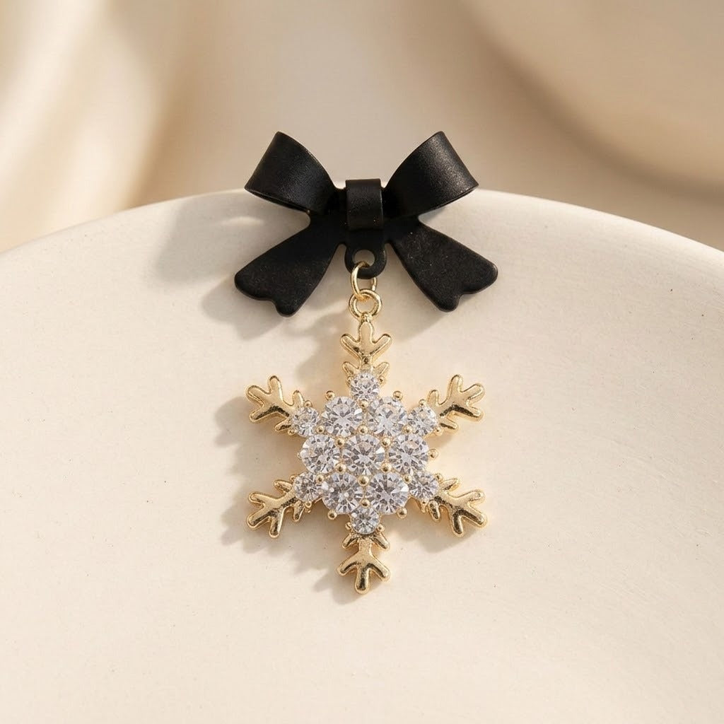 A black bow earring with a gold-tone snowflake drop and clear stones rests on a curved cream display surface.