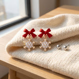 Red-bow snowflake drop earrings with clear rhinestones and gold-colored outlines rest on cream knit beside silver-colored ball studs near a window.