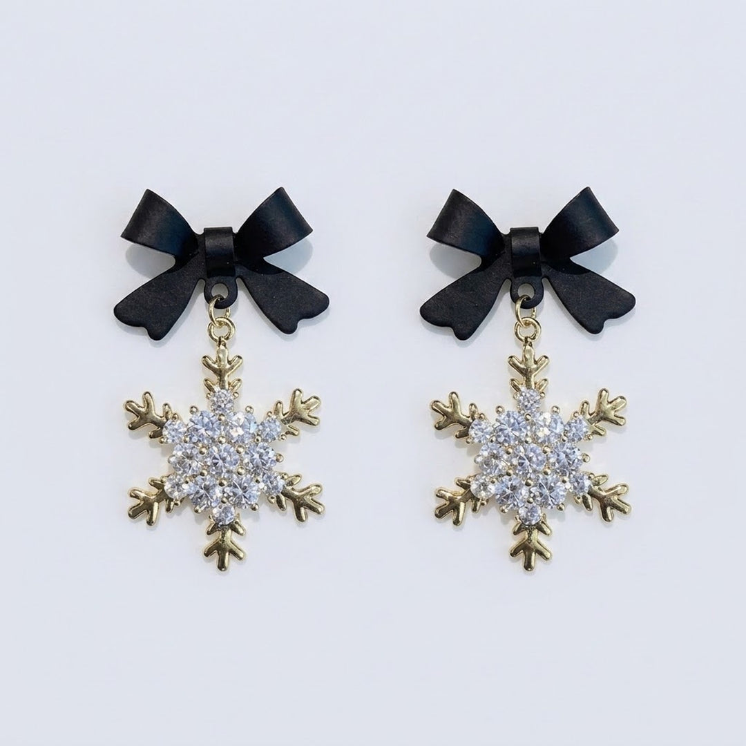 Snowflake drop earrings with black bow tops, clear rhinestones and gold-colored outlines lie side by side on a pale background.