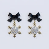 Snowflake drop earrings with black bow tops, clear rhinestones and gold-colored outlines lie side by side on a pale background.