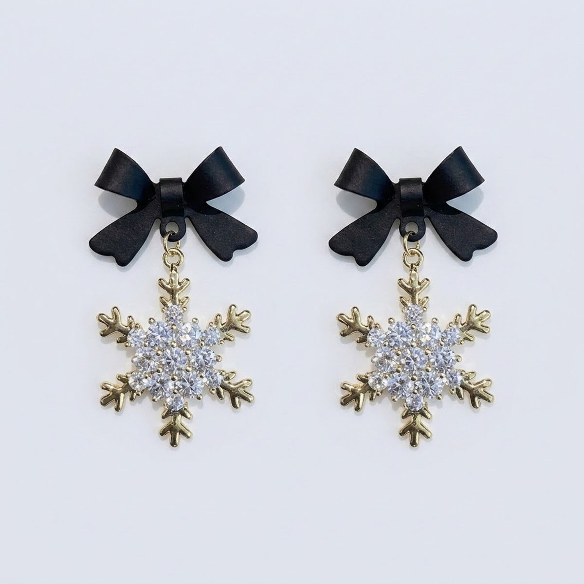 Snowflake drop earrings with black bow tops, clear rhinestones and gold-colored outlines lie side by side on a pale background.