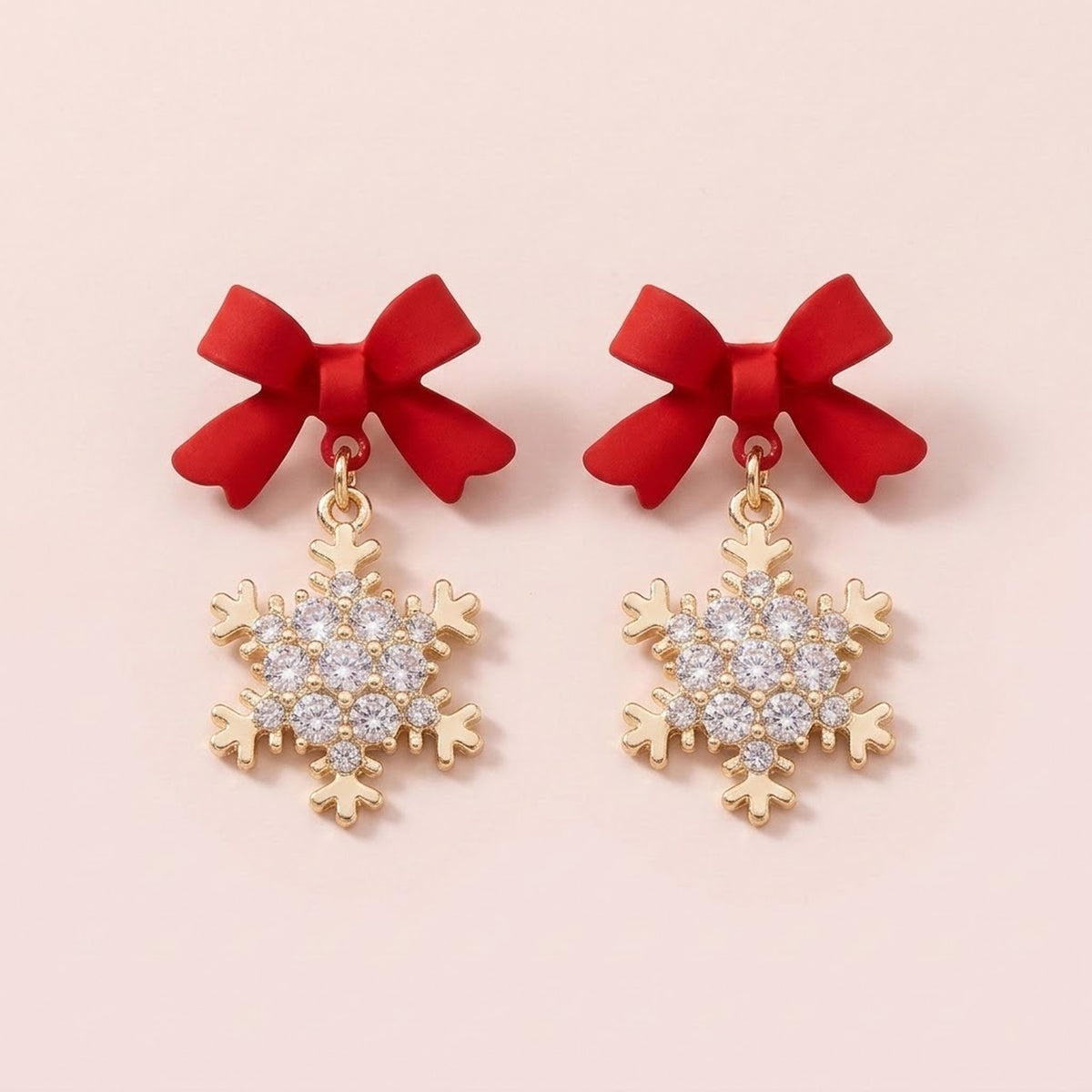 A pair of red bow earrings with gold-tone snowflake drops and clear stones sits against a pale pink background.