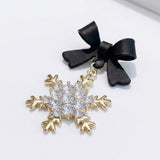 A black bow earring lies diagonally on a white surface, showing its gold-tone snowflake drop and raised clear stones.
