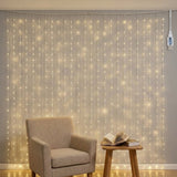 Warm white curtain lights hang against a gray wall behind a beige armchair and wooden table with books.