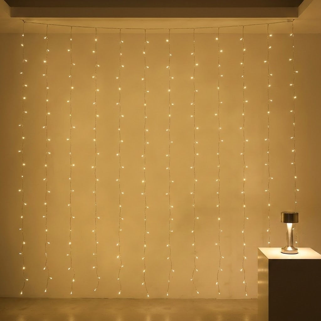 Warm white curtain lights hang against a cream wall beside a small table holding a metallic lamp.