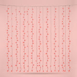 Red curtain string lights hang in vertical strands from a horizontal wire against a pale pink background.