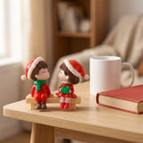 Two small Christmas-themed figurines sitting on a wooden table, styled with a white mug and a red book in a warm, softly blurred indoor setting.