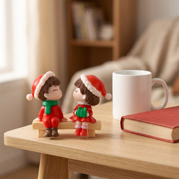 Two small Christmas-themed figurines sitting on a wooden table, styled with a white mug and a red book in a warm, softly blurred indoor setting.