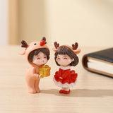 Plastic Christmas figurines in a peach reindeer outfit and red dress stand on a pale wooden surface beside a dark book.